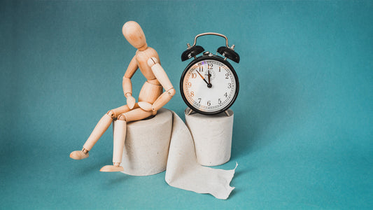  Do Iron Pills Cause Constipation? A wooden anatomical figure is sat on top of a toilet roll holding its stomach. Next to it sits a clock. Supporting the idea of constipation.