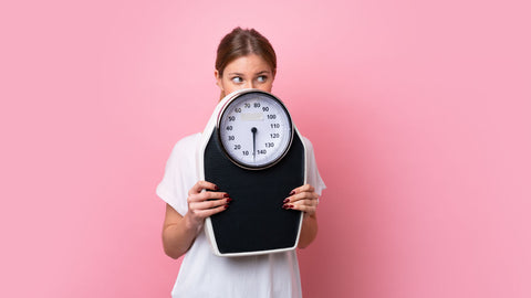 Woman hiding behind bodyweight scales, poses the question, does biotin cause weight gain?