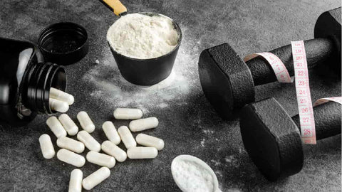 Creatine Before or After Workout: When Should You Take It? Creatine supplements as white powder in scoops and white capsules spilling out of a bottle. Next to them sits a pair of dumbbells with a measuring tape draped over them.